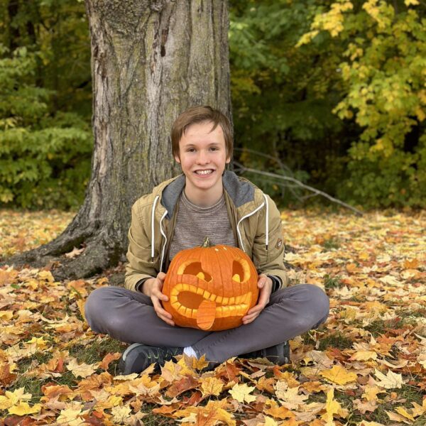 An exchange student sitting under a tree holding a carved pumpkin.