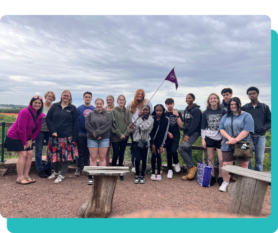 A group of teens with their chaperone smiling for a group photo. One student is holding a purple YFU flag.