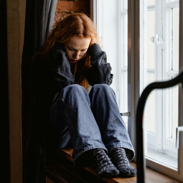 A girl holds her head while sitting by a window. She looks sad.